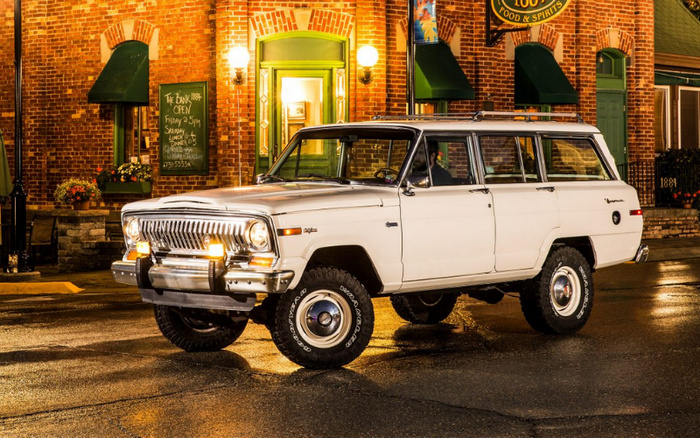 Classic white Jeep Wagoneer parked on a wet street in front of a brick building with green trim and lit lamps at night. The building has a sign that reads 'The Drink Room OPEN Friday 3-5PM, Saturday 1-Lonely J Dinners 4-9, Sunday Closed, 270-555-3454'. The scene is well-lit with warm street lights reflecting on the wet pavement. The image depicts a vintage used car in an urban setting, suitable for advertising used cars or new cars.