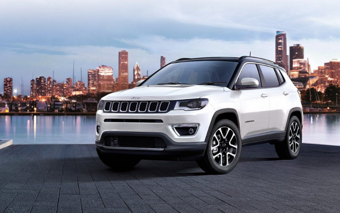White Jeep Compass SUV parked on a dark paved surface with a city skyline and waterfront in the background during twilight, representing used cars and new cars available for sale.