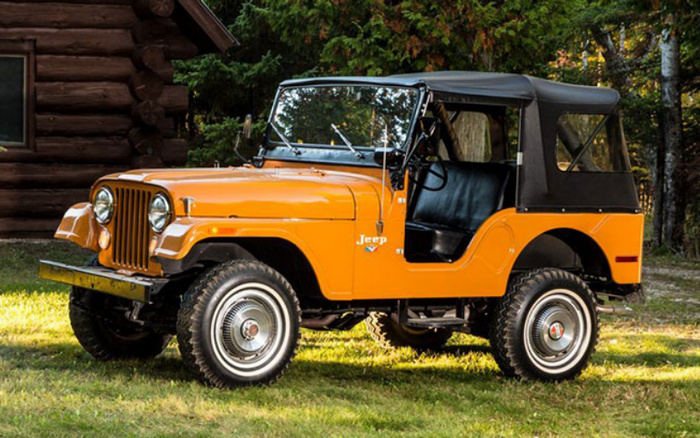 Orange classic Jeep with black soft top parked on grass in front of a wooden cabin and trees, showing side profile with visible large off-road tires and Jeep logo on the side, ideal for used cars and new cars enthusiasts.