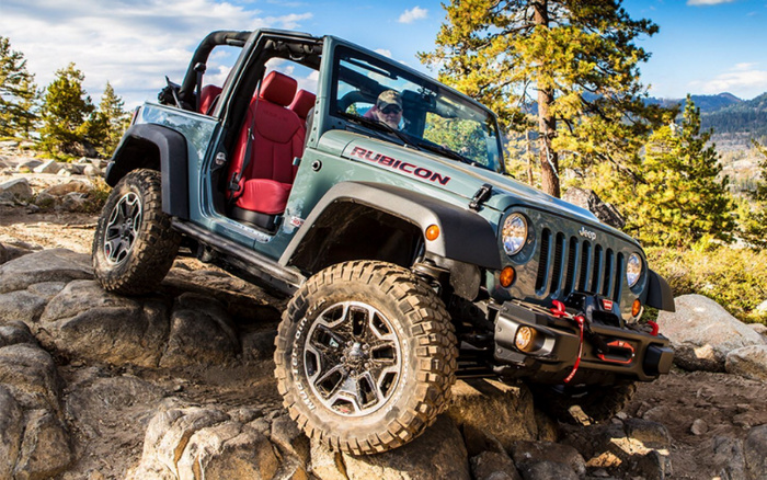 Green Jeep Rubicon with open doors and red interior seats climbing over rocky terrain in a mountainous forest area with pine trees and clear blue sky, showcasing off-road capabilities of used cars and new cars.