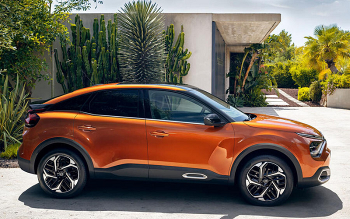 Side view of an orange modern crossover car parked in front of a contemporary house surrounded by lush green plants and palm trees, showcasing a sleek design with black tinted windows and black trim around the wheel arches and lower body panels, representing options available in used cars and new cars markets.