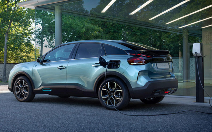 Light blue electric car plugged into a charging station outside a modern glass building with trees reflected in the windows, illustrating new cars with electric technology, suitable for used cars and new cars market.