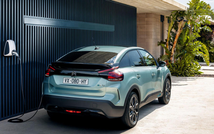A light blue Citroën electric vehicle parked and charging at a modern home with a dark blue slatted wall and leafy green plants nearby. The car's licence plate reads VX-080-HY. This image features a new car model, highlighting clean design and eco-friendly electric charging, suitable for advertisements or listings involving used cars and new cars.