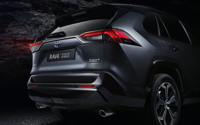 Rear view of a dark grey Toyota RAV4 Plug-in Hybrid AWD vehicle against a dark stone wall background, showing the rear lights, dual exhaust pipes, and badges including 'RAV4 Plug-in Hybrid' and 'AWD'.