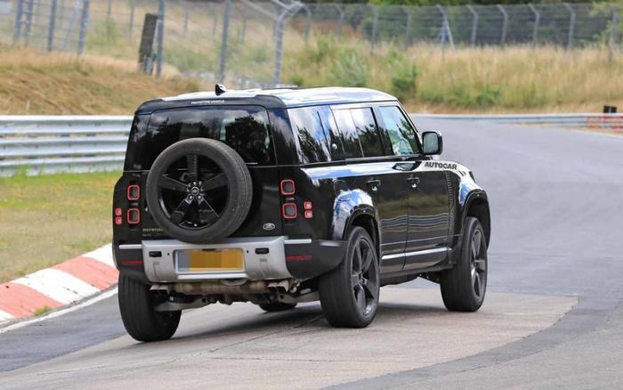 Black Land Rover Defender prototype vehicle with a spare wheel mounted on the rear door driving on a race track with safety barriers and grassy surroundings, featuring the text 'PROTOTYPE VEHICLE' on the rear top and 'AUTOCAR' on the side mirror, suitable for illustrating used cars and new cars.