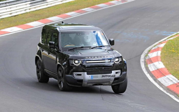 Black Land Rover Defender driving on a race track with red and white curbs, front view showing the word 'DEFENDER' on the hood and a license plate with the text 'AUTOCAR'. The image represents both used cars and new cars in the context of automotive testing and review.