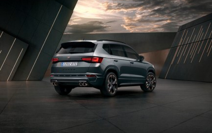 Rear view of a grey Cupra Ateca SUV parked indoors under modern architectural lighting. The car has distinctive quad exhaust pipes, sporty alloy wheels, and a license plate reading 2202 ATG. The scene features a dramatic, cloudy sky visible through an opening, emphasizing the vehicle's stylish and modern design. Ideal for showcasing used cars and new cars in a contemporary setting.