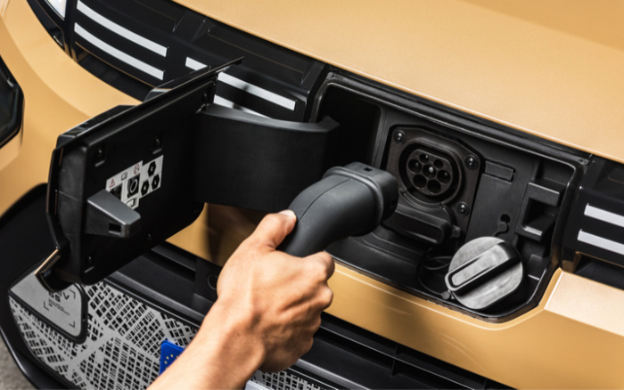 Close-up of a hand plugging an electric vehicle charging connector into a sandy gold-coloured car's front charging port, highlighting the electric charging feature for used cars and new cars.