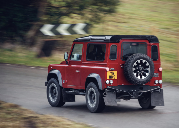 Red Land Rover Defender 90 with black roof and spare tire mounted on the rear door driving on a curved rural road with chevron road signs in the background, showcasing a classic used car model alongside new cars options.