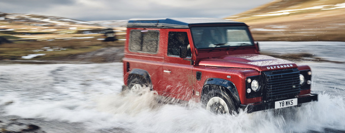Red Land Rover Defender driving through a shallow river in a rugged outdoor setting with hills in the background, showcasing its off-road capability. The vehicle has a white and black UK license plate reading Y8 WKS. Suitable for used cars and new cars buyers looking for durable and versatile 4x4 vehicles.