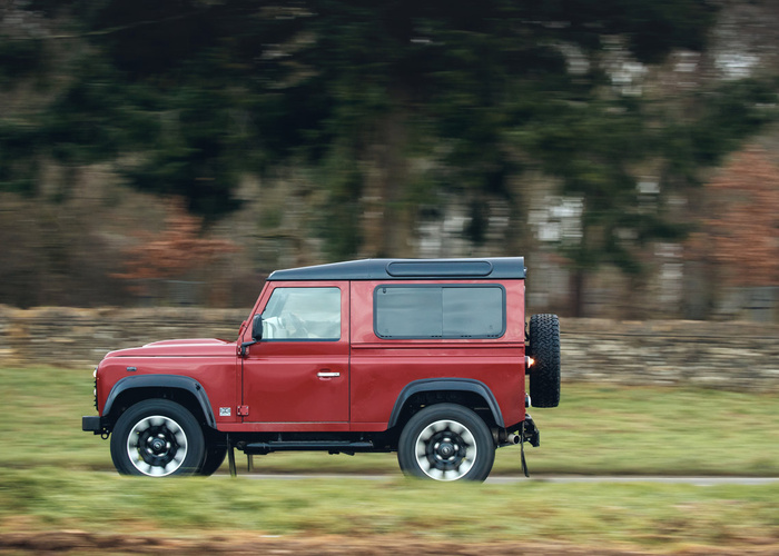 Red Land Rover Defender 90 with black roof driving past stone wall and trees on a rural road, representing sturdy used cars and new cars for off-road and everyday driving.