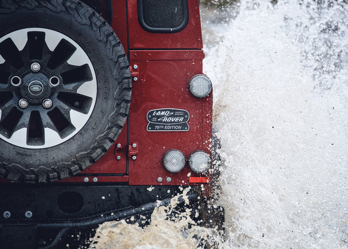 Close-up of the rear side of a red Land Rover 70th Edition vehicle splashing through water, showing the spare tyre with Land Rover logo, rear lights, and the badge indicating the 70th Edition and Solihull, England marking, highlighting rugged used cars and new cars capabilities.