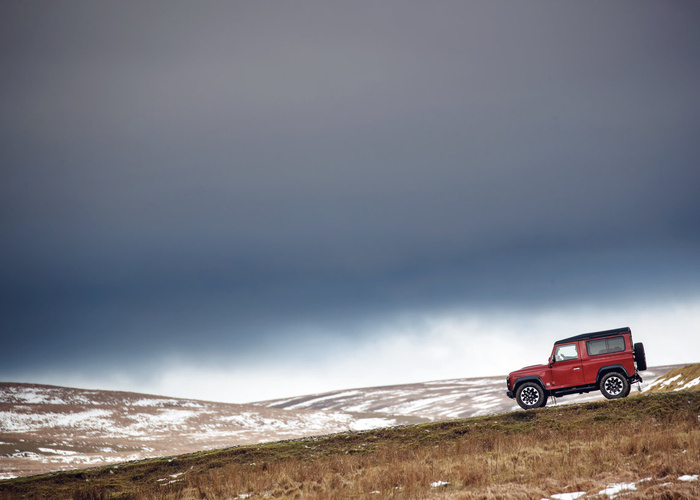 Red 4x4 off-road vehicle driving on a grassy and snowy hillside under a dark, cloudy sky with rolling hills in the background, showcasing an example of used cars and new cars suitable for rugged terrain.