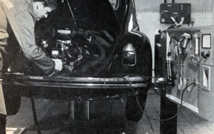 Black and white vintage photo showing a man working on the engine of a car with the bonnet open. The car is elevated on a mechanical testing rig in a workshop with various diagnostic tools and gauges on the wall. The scene highlights historical vehicle inspection or repair, relevant to both used cars and new cars maintenance and testing.