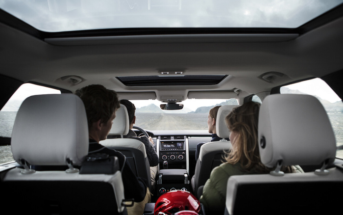 Interior view from the back seat of a car with four passengers driving on a gravel road in a mountainous area, showing the dashboard, steering wheel, panoramic sunroof, and a red helmet on the rear seat. Suitable for keywords used cars, new cars.