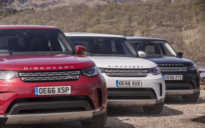 Front view of three Land Rover Discovery cars parked outdoors on gravel with a natural backdrop of trees and hills; vehicles shown in red, white, and black colours, each with UK registration plates OE66 XSP, OE66 XUX, and OE66 XTS respectively; image suitable for used cars and new cars listings or promotions.