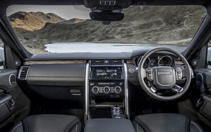 Interior view of a modern luxury car from the driver's perspective showing the steering wheel, dashboard with touchscreen display, climate control dials, and leather seats. The car is parked by a scenic lake with hills in the background. Keywords used cars, new cars.