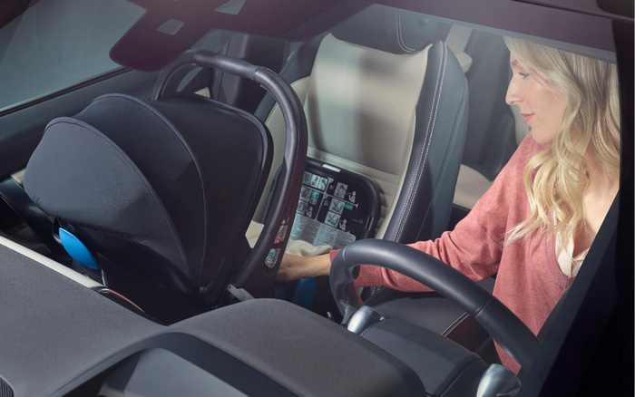 Interior view of a modern car with a woman installing a black infant car seat in the back, visible through the front windshield. The focus is on the car seat and the woman securing it while seated in the driver's seat, showcasing safety and family orientation in used cars and new cars.