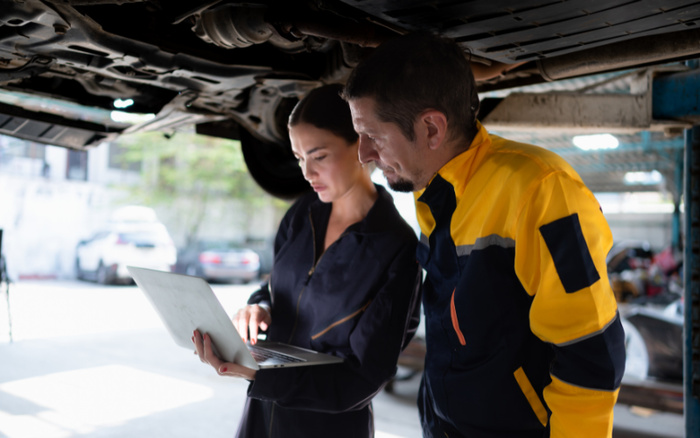 A male and female technician standing under a suspended vehicle reviewing a laptop screen 