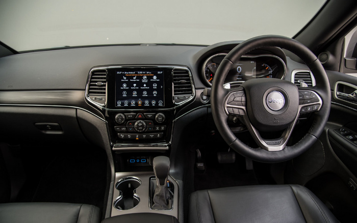 Interior view of a Jeep car showing the dashboard, steering wheel with Jeep logo, touchscreen infotainment system displaying radio, media, climate, and vehicle controls, gear shift lever, cup holders, and black leather seats, highlighting features relevant to buyers of used cars and new cars.