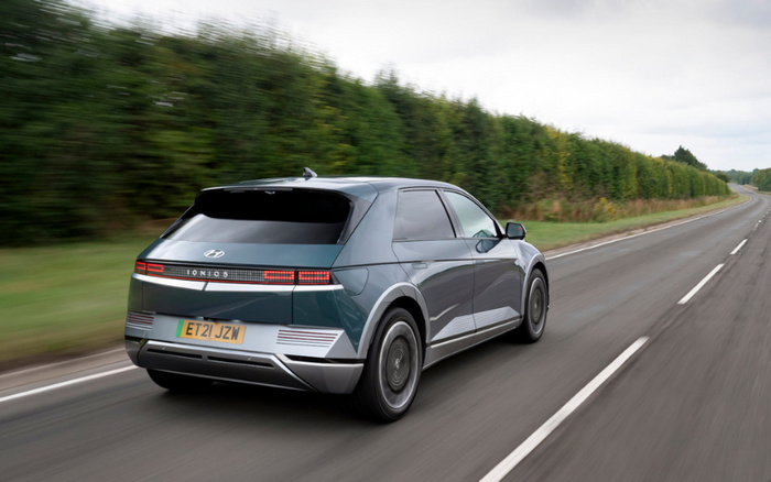 Rear view of a green Hyundai Ioniq 5 electric car driving on a country road with a hedge alongside. The car's UK license plate reads ET21 JZW. The image represents new cars, with a focus on modern electric vehicles, suitable for searches related to used cars and new cars.