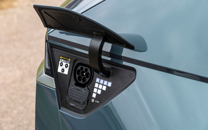Close-up of an electric car charging port with the cover open, showing the Type 2 connector and warning labels on the inside panel. The car is a sleek metallic blue-grey colour, highlighting the modern design of new cars and the advancing technology in used cars.