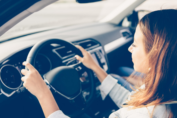 Woman sitting in the driver's seat of a car holding the steering wheel with both hands, viewed from the side. The modern car interior includes a dashboard with various controls and screens visible, representing used cars and new cars for sale.