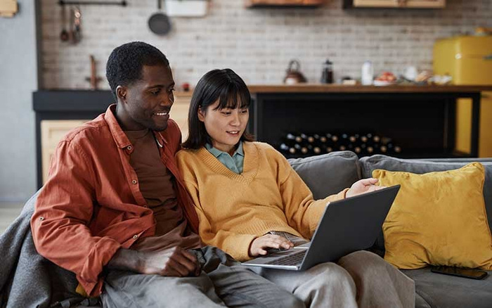 Young couple sitting together on a grey couch in a modern living room, looking at a laptop screen with interest. The man is wearing a rust-coloured jacket over a brown shirt, and the woman is dressed in a mustard-yellow sweater over a blue collared shirt. The room features mustard-yellow pillows, a dark sideboard with wine bottles stored underneath, and light brick walls with kitchen utensils hanging. The image suggests browsing or shopping, potentially for used cars or new cars online.