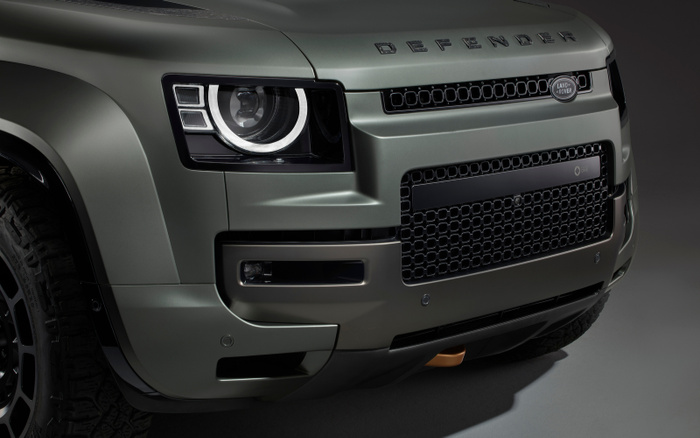 Close-up view of the front of a Land Rover Defender in metallic grey, showcasing the distinctive Defender lettering on the bonnet, the Land Rover badge on the grille, LED headlights, and rugged tire, representing both used cars and new cars.