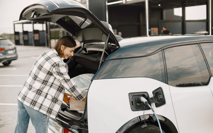 A woman in a checkered jacket and jeans loading a reusable shopping bag into the open trunk of a white electric car charging at a station, representing modern used cars and new cars with eco-friendly features in a bright, clean dealership or parking area.