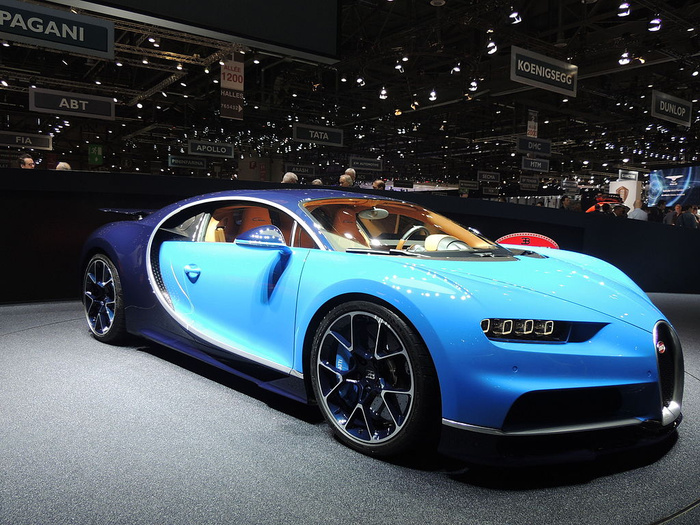 A shiny blue Bugatti sports car with tan leather interior displayed at a car show surrounded by signs for luxury and performance car brands such as Pagani, Koenigsegg, Dunlop, ABT, and Apollo. The setting is an indoor exhibition hall filled with bright lights and people in the background. This scene showcases new cars, capturing the sleek design and premium features typical of luxury used cars and new cars available in the high-end automotive market.