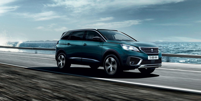 Dark teal Peugeot SUV driving on a coastal highway with the ocean and waves in the background, clear sky above, vehicle has license plate TX-080-WH. Image suitable for used cars and new cars showcasing a modern family car in motion.
