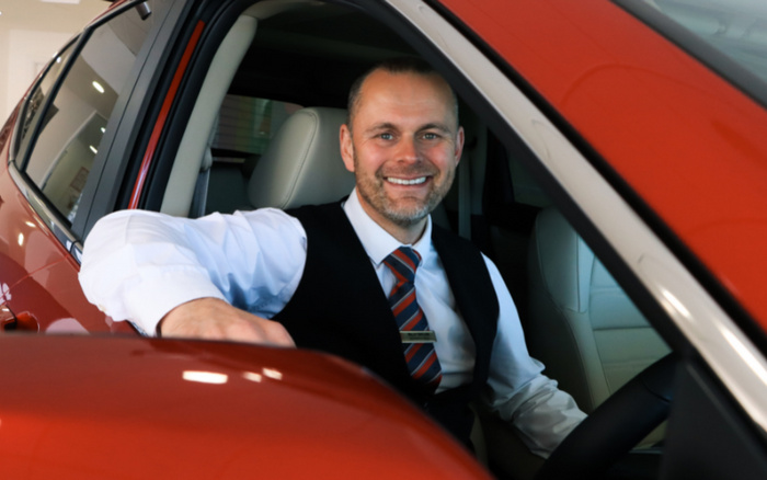 Smiling male car salesman sitting in the driver's seat of a shiny red car, wearing a white shirt, black waistcoat, and a red and blue striped tie, representing a dealership that sells new cars and used cars.