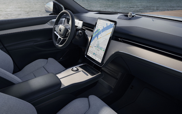 Interior of a modern car featuring a sleek dashboard with a large vertical touchscreen displaying a navigation map. The steering wheel has a minimalist design with the Volvo logo in the centre. The seats are upholstered in grey fabric, and the centre console has a wood-finish panel with a rotary dial and storage compartments. The car is parked near a body of water, visible through the window. This image represents the interior features that can appeal to buyers of used cars and new cars.