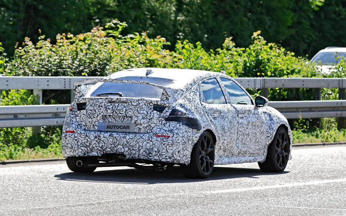 Prototype car covered in white and black swirling camouflage driving on a road next to a metal guardrail and greenery, with a rear license plate displaying the text AUTOCAR, representing a new car model undergoing testing, relevant for both used cars and new cars markets.