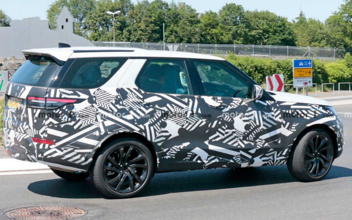 Side view of a new car prototype covered in black and white camouflage wrap driving on a road with greenery and road signs in the background. The car, featuring matte black wheels and a spoiler, is undergoing testing and is likely a new car model being evaluated before release. The scene suggests the car is part of new cars development, relevant to used cars and new cars market discussions.