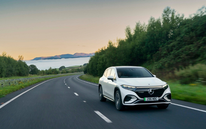 White Mercedes-Benz electric car driving on a curved countryside road with greenery and trees on both sides, clear sky with mountain range in the background, license plate 296 MBC. Image suitable for used cars and new cars.