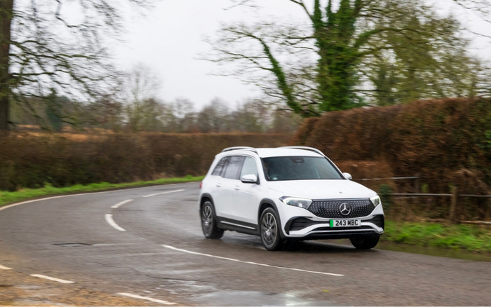 White Mercedes-Benz electric SUV driving on a winding country road with licence plate 243 MBC, surrounded by bare trees and hedges, representing both used cars and new cars.