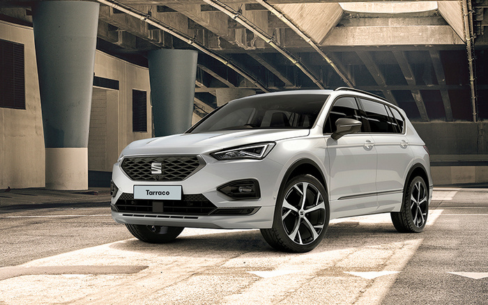 White SEAT Tarraco SUV parked under a concrete bridge structure with sunlight casting shadows. The vehicle is a modern model with sleek LED headlights and large alloy wheels, suitable for both used cars and new cars buyers.