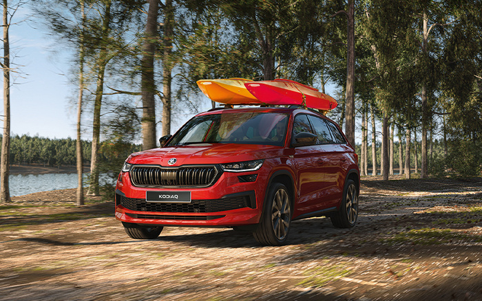 Red Škoda Kodiaq SUV parked on a forest path with two kayaks, one red and one yellow, secured on its roof rack. The image highlights a new car model surrounded by trees near a lake, emphasising outdoor adventure and family travel. Suitable for used cars and new cars searches.