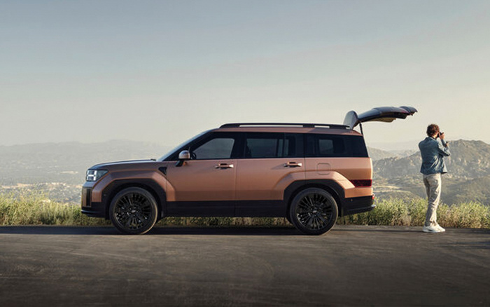 Side view of a modern bronze and black SUV parked on a roadside with its rear hatch open, a person standing near the open trunk taking photos with a camera, mountainous landscape in the background, showcasing a new car model suitable for outdoor adventures, ideal for buyers interested in used cars and new cars.