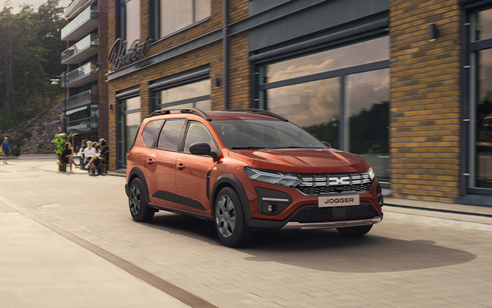 Orange Dacia Jogger car driving on a paved street next to a modern building with large windows and brick walls. The car has a front license plate reading 'JOGGER'. Several people are sitting and walking on the sidewalk in the background. Image suitable for used cars and new cars listings.