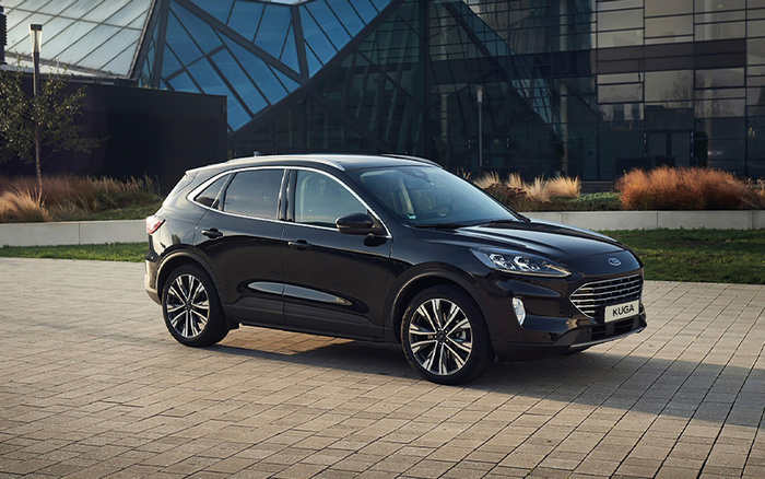 Black Ford Kuga parked on a tiled pavement in front of a modern glass building, showcasing the sleek design and alloy wheels of the new model, ideal for those seeking new cars or used cars in excellent condition.