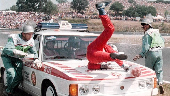 A dramatic scene at a car racing event showing a marshal in red overalls being thrown onto the bonnet of a white emergency vehicle marked 'ASSISTANCE C 124'. Two other marshals in green uniforms and helmets are attending to the situation. The background includes a large crowd of spectators. The vehicle bears logos and text indicating association with a motorsport club. Keywords used cars, new cars.