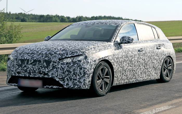 A camouflaged prototype car driving on a road with green fields and a wind turbine in the background, showcasing a hatchback design covered in a geometric grey and black pattern used for new cars testing before release, relevant for used cars and new cars market.