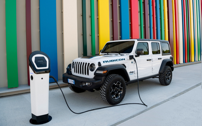 White Jeep Rubicon electric vehicle plugged into a charging station outside a building with vertical multicoloured panels in green, red, blue, yellow, and pink shades, illustrating modern new cars and eco-friendly used cars.