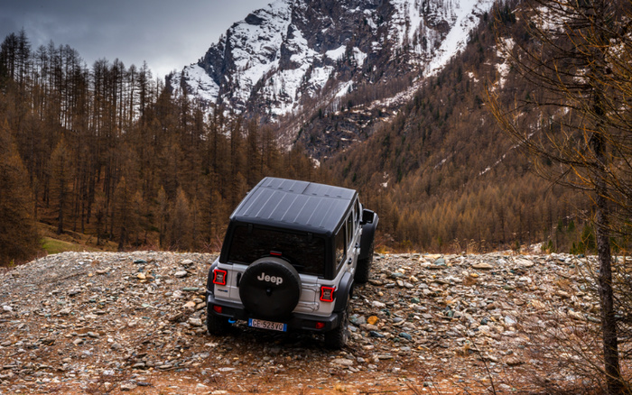 Silver Jeep off-road vehicle driving on a rocky trail in a mountainous forest area with snow-covered peaks in the background. The Jeep is facing away, displaying a spare tyre cover with the Jeep logo. The image evokes adventure and rugged terrain, suitable for promoting used cars and new cars with off-road capabilities.