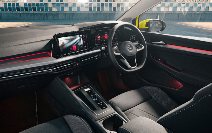 Interior of a modern Volkswagen car showing the driver's seat, steering wheel with VW logo, and digital dashboard with two screens displaying vehicle and rear camera views. The car features black fabric seats with patterned inserts, a sleek centre console with gear selector, cup holders, and door panel with integrated controls. The interior has ambient red lighting along the dashboard and door, set against a backdrop of a tiled wall and yellow side mirror visible outside. Ideal for showcasing used cars and new cars with advanced technology and stylish design.