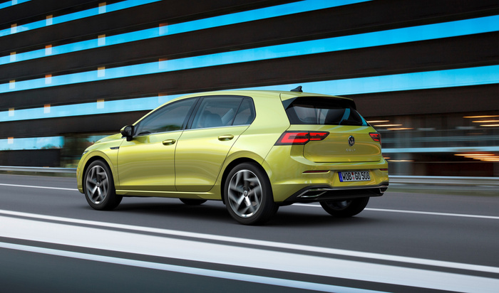 Light green Volkswagen Golf hatchback driving on a road with five doors and tinted windows, showing rear and side view. The car has a German licence plate reading 'WOB GO 508' and modern LED tail lights. The background features a building with horizontal blue and brown stripes, captured in motion blur. Ideal image for used cars and new cars dealership or sales.
