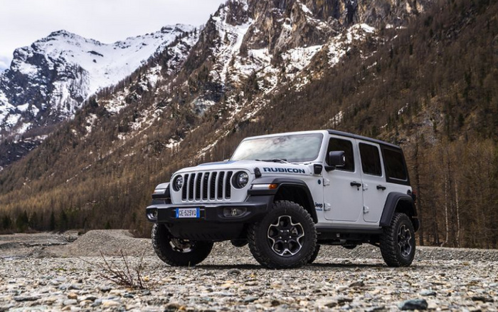 White Jeep Wrangler Rubicon 4-door off-road vehicle parked on a rocky ground with snow-capped mountain range and dense forest in the background, featuring a European license plate CE-525VO, showcasing rugged all-terrain tyres and black accents, suitable for advertising used cars and new cars in adventurous and durable vehicle categories.